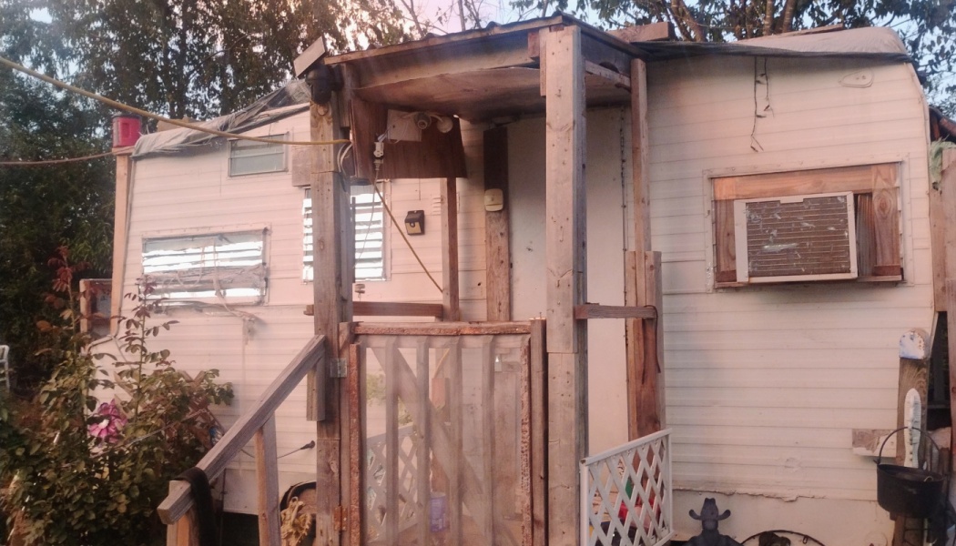 A weathered white mobile home with a makeshift wooden porch structure and an exterior window air conditioner.
