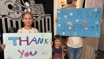 Smiling children hold colorful, handmade signs reading "Thank You" and "FOR UR SUPPORT."