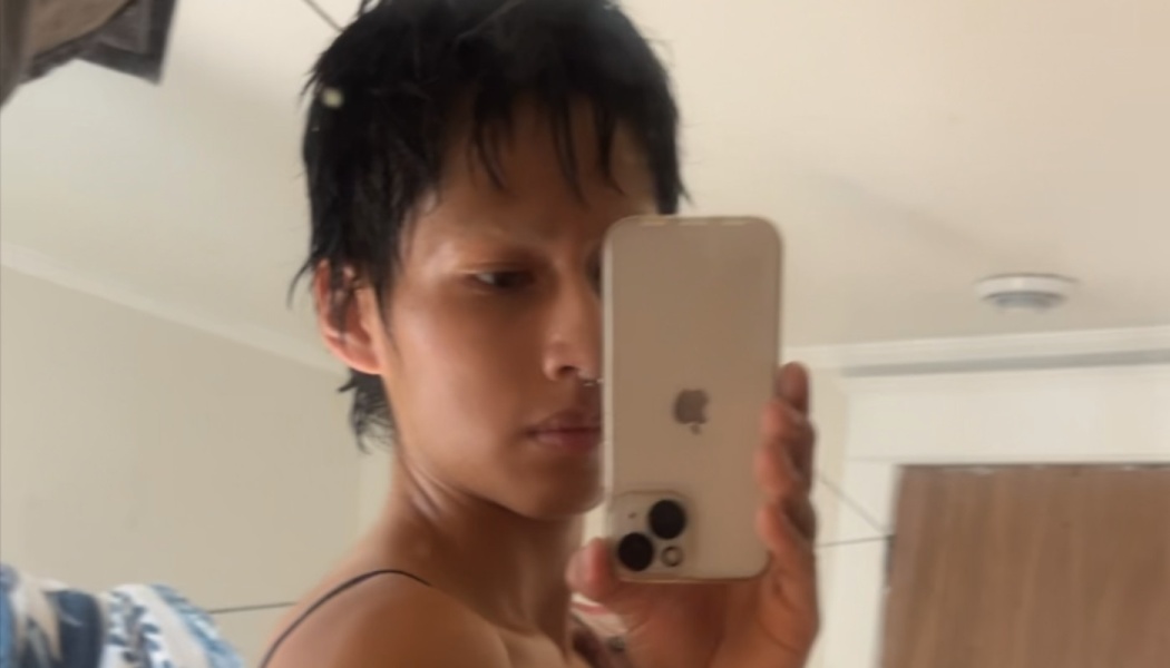Person with short dark hair taking a mirror selfie using a smartphone indoors.