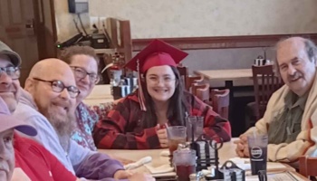 A smiling graduate in a red cap sits at a restaurant table surrounded by her family and friends.