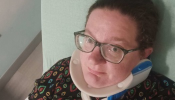A person with glasses wearing a medical neck brace lying down in a hospital bed.