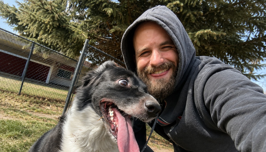 A smiling bearded man in a hoodie takes a selfie with a silly black and white dog sticking its long tongue out.