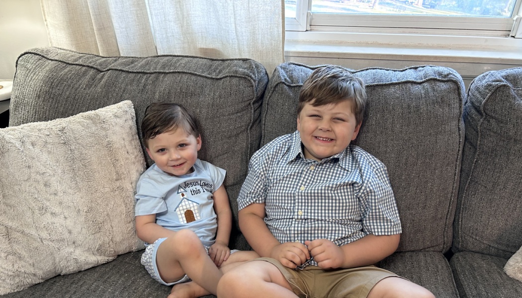 Two young brothers sitting together on a grey couch, smiling happily at the camera.