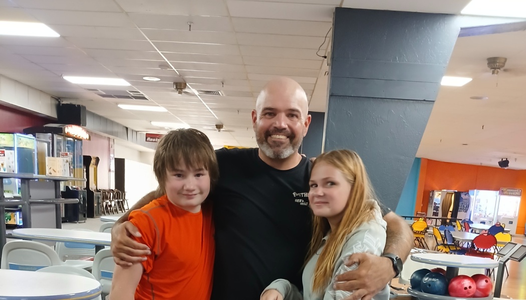 A smiling bald man hugs a young boy in an orange shirt and a young girl at a bowling alley.