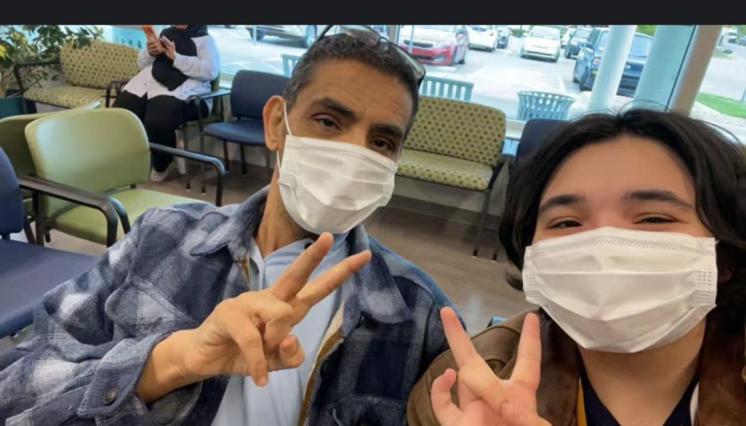 A man in a blue plaid jacket and a young companion wearing face masks make peace signs in a medical waiting room.