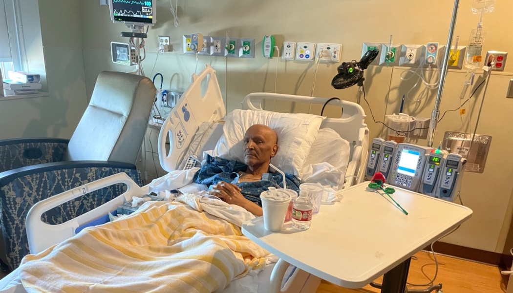 An elderly man rests in a hospital bed surrounded by medical equipment, IV pumps, and monitors.