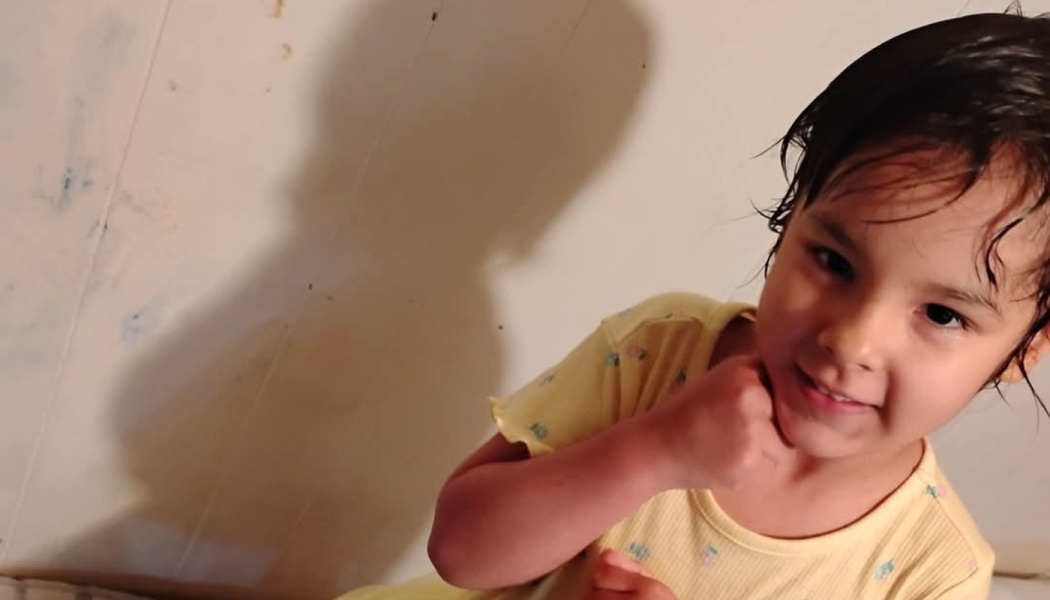 A smiling young girl with dark hair wearing a yellow shirt, resting her hand under her chin.