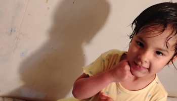 A smiling young girl with dark hair wearing a yellow shirt, resting her hand under her chin.