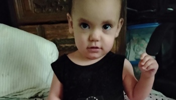 A toddler with dark eyes wearing a black dress looking directly at the camera with a raised hand.
