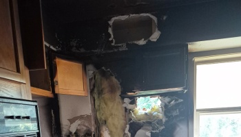 Kitchen severely damaged by fire with charred walls, soot-covered cabinets, and holes exposing insulation.
