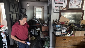 A man in a red shirt holds his phone inside a cluttered, smoke- and soot-damaged room after a fire.