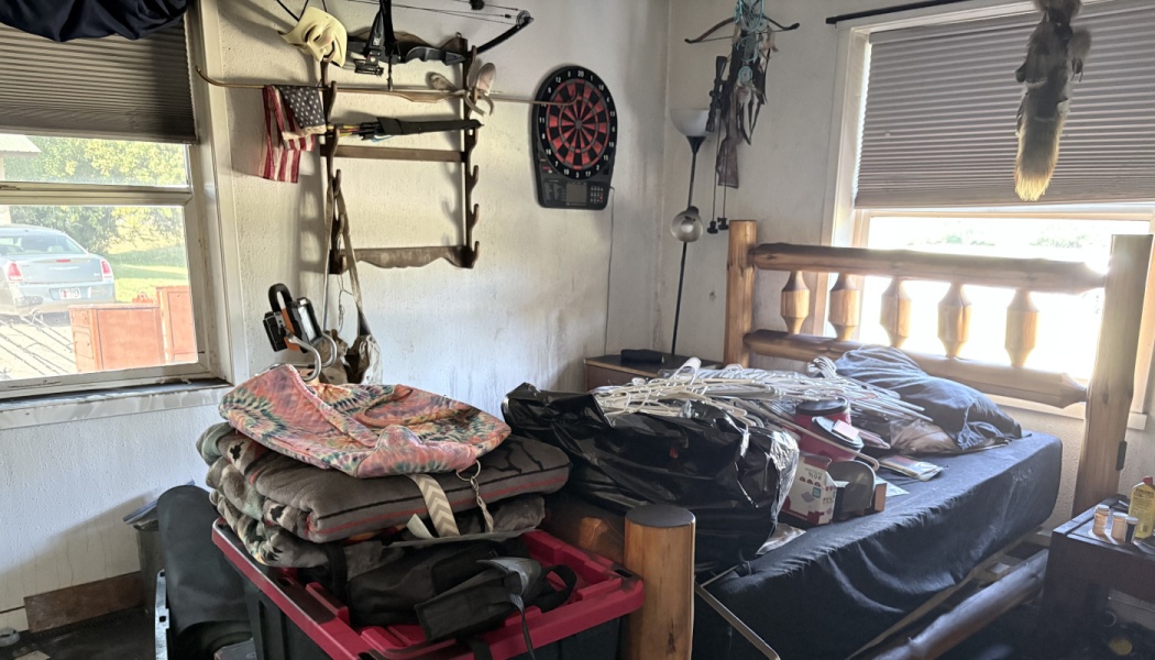 A cluttered bedroom with stacked blankets, plastic hangers, and bags piled on a log bed and storage bin.