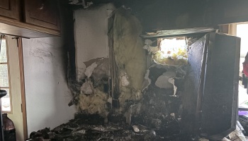 Severely fire-damaged kitchen wall with charred drywall, exposed yellow insulation, soot, and burnt appliances.