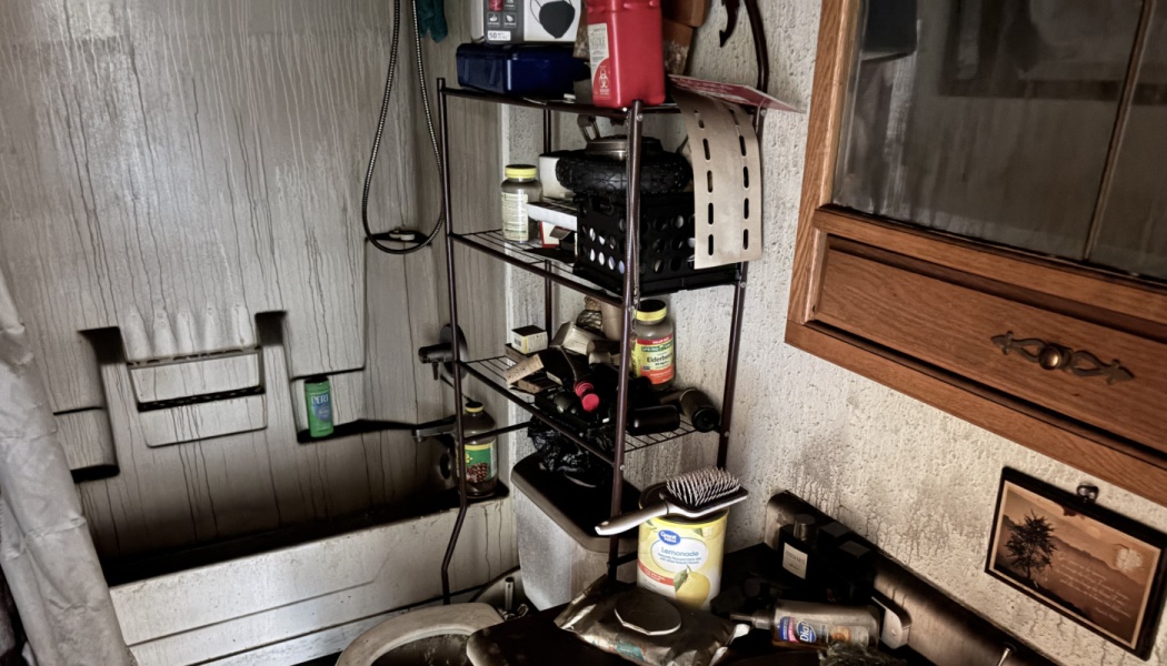 Fire-damaged bathroom with heavy soot coating the walls, shower tub, toilet, and cluttered storage shelves.
