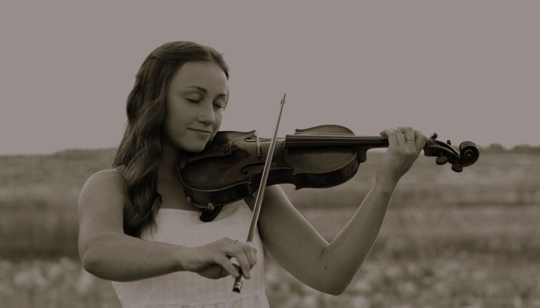 A young woman playing the violin with her eyes closed outdoors, captured in a soft sepia tone.