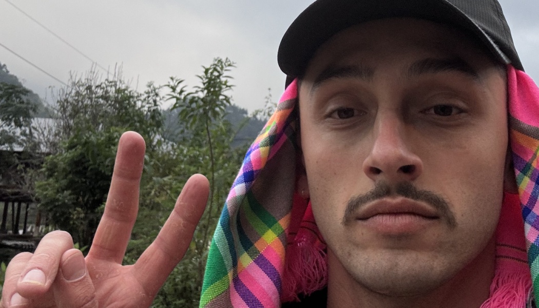 Close-up selfie of a man with a mustache making a peace sign, wearing a cap over a colorful patterned scarf.
