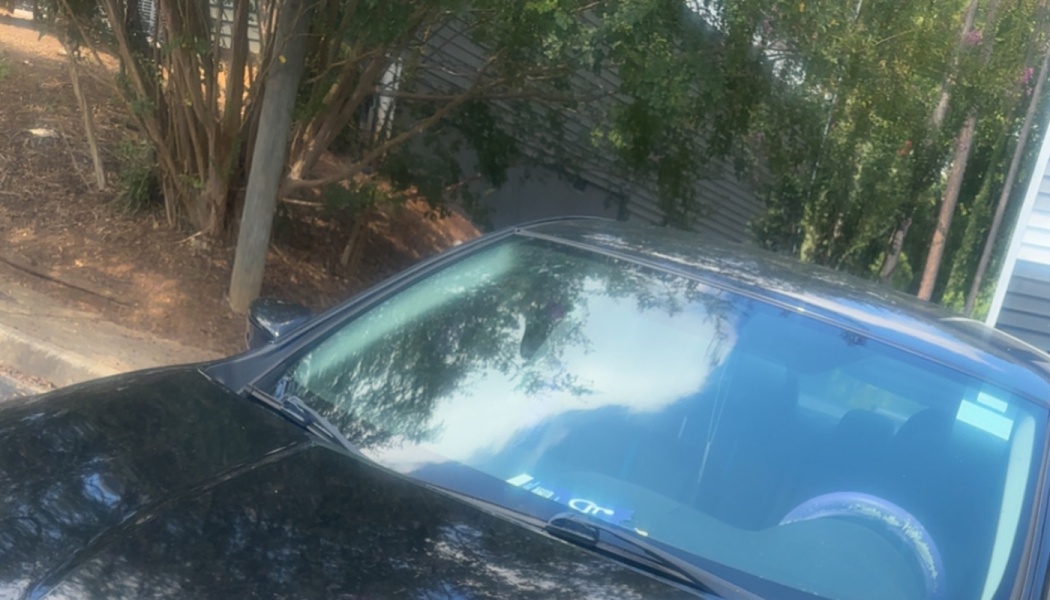 Front view of a dark car's hood and windshield reflecting green trees and sky outdoors.