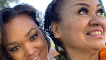 Two smiling women posing close together, wearing traditional leis and floral hair accessories.