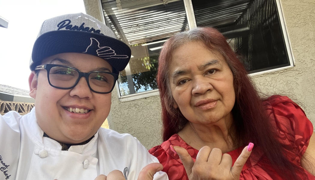 A smiling chef wearing a cap poses with an older woman in red, both making shaka hand gestures outdoors.