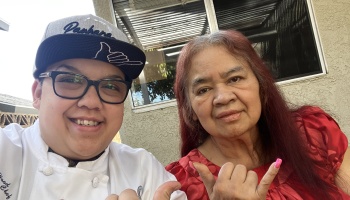 A smiling chef wearing a cap poses with an older woman in red, both making shaka hand gestures outdoors.