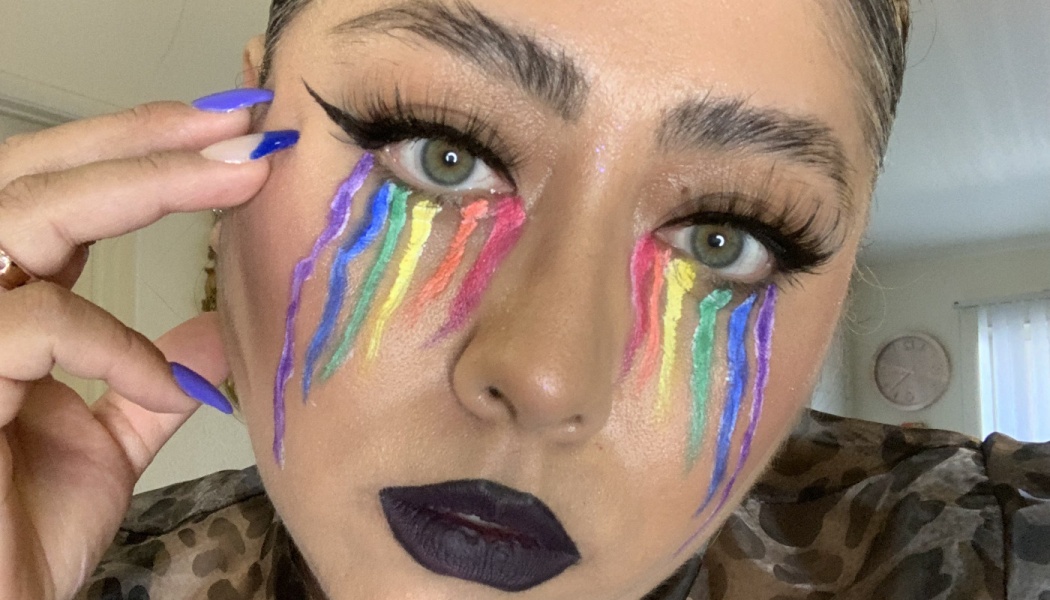 Person with rainbow-painted tears streaming from their eyes, dark purple lipstick, and sharp winged eyeliner.