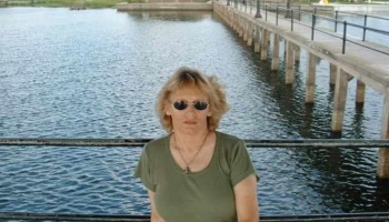 Blonde woman wearing sunglasses and a green t-shirt standing near water with a pier in the background.