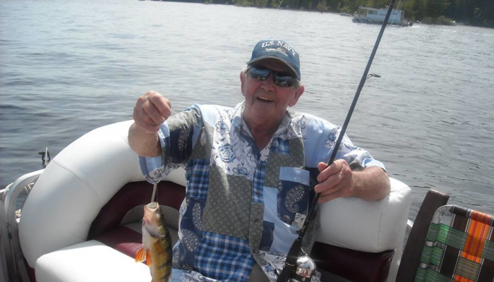 A smiling elderly man in a U.S. Navy cap sits on a boat, proudly holding up a small fish he caught.