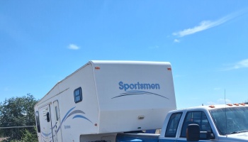 A white Sportsmen fifth-wheel camper trailer hitched to a pickup truck under a clear blue sky.