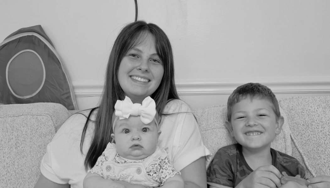 A smiling mother sitting on a couch holds her baby wearing a bow headband, next to her smiling young son.