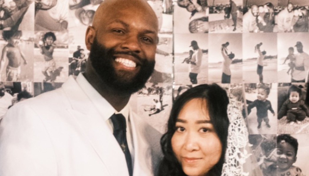 A smiling groom in a white suit and bride in a lace veil posing in front of a family photo collage.