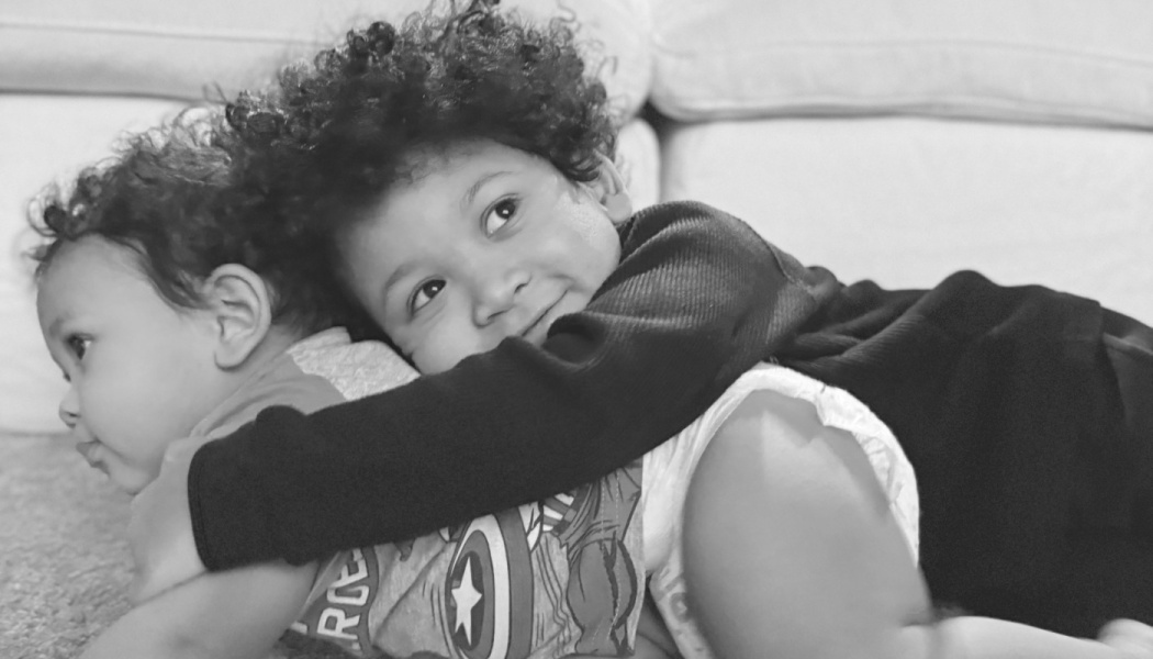 A smiling young boy with curly hair hugs his younger sibling affectionately from behind.