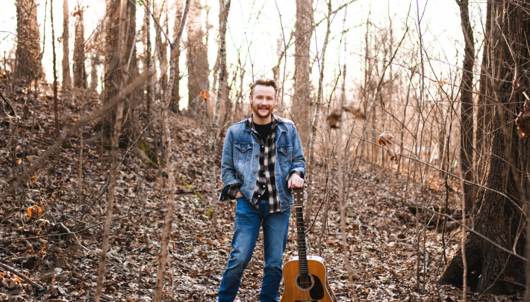 A smiling musician in a denim jacket stands in a wooded forest, resting his hand on an acoustic guitar.