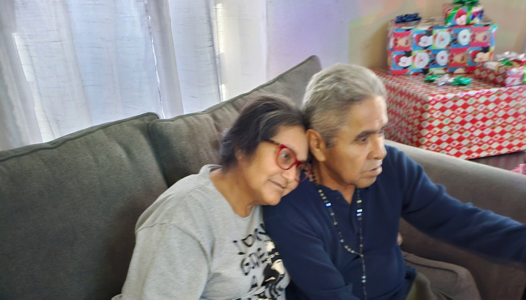 An elderly woman with red glasses leans her head affectionately on a man's shoulder as they sit on a couch.
