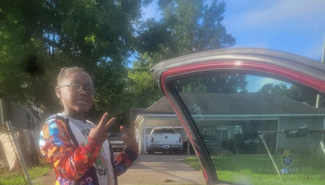 Young girl wearing glasses and a colorful jacket posing with peace signs next to an open car door outside.