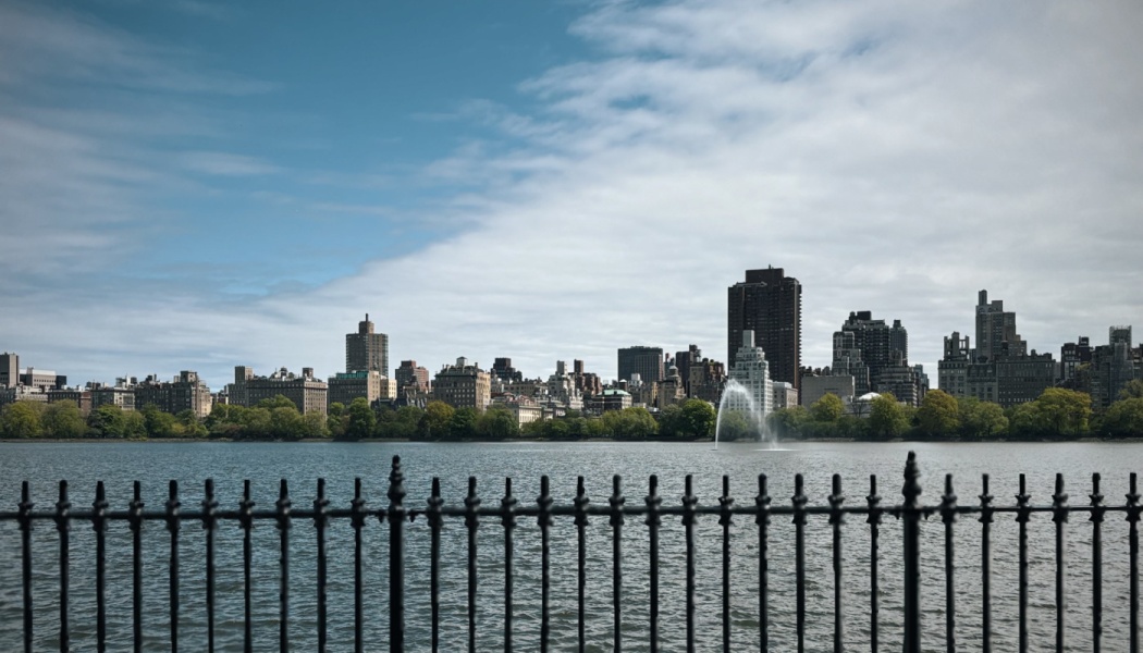 Black wrought-iron fence overlooking a wide reservoir, with a city skyline and fountain across the water.