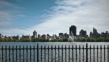 Black wrought-iron fence overlooking a wide reservoir, with a city skyline and fountain across the water.