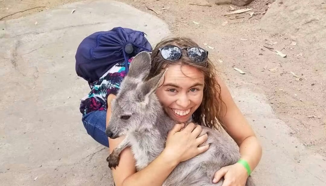 A smiling woman with a backpack gently hugs a young kangaroo joey.