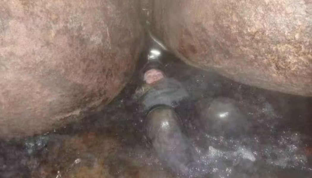 A caver floating in water, squeezing through an extremely narrow rock crevice inside a dark cave.