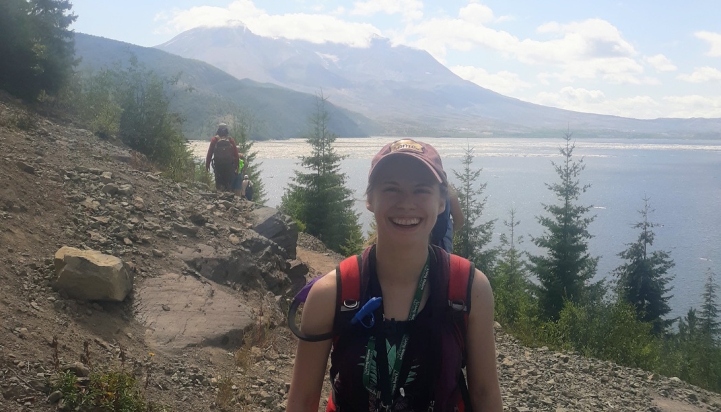 A smiling young woman with a backpack hikes a rocky trail overlooking a large lake and mountain peak.