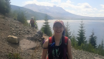 A smiling young woman with a backpack hikes a rocky trail overlooking a large lake and mountain peak.