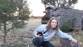A smiling young woman with a camera crouches outdoors, petting a wombat next to a large fallen log.
