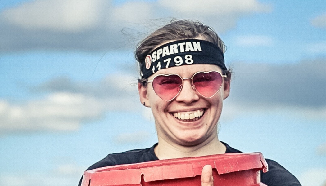 A smiling woman wearing a Spartan headband and pink heart-shaped sunglasses, holding a red bucket.