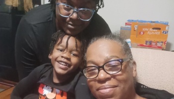 A smiling young Black boy, an adult, and an older woman with glasses posing closely together indoors.