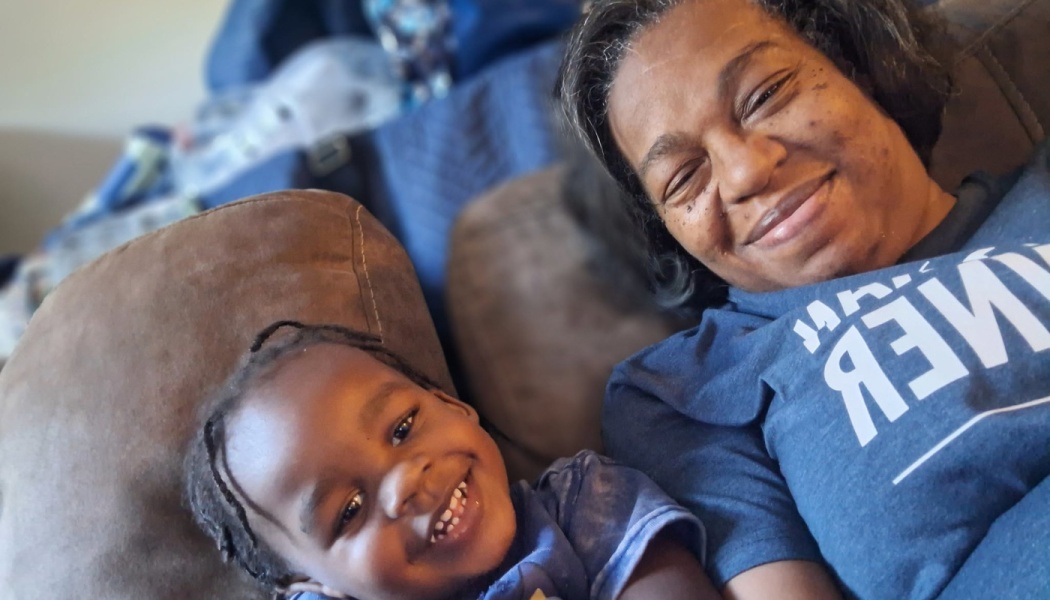 A smiling woman and young child lie on a couch together, looking happily at the camera.