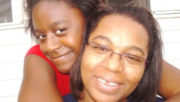 A smiling young Black girl hugging a smiling Black woman with glasses from behind.