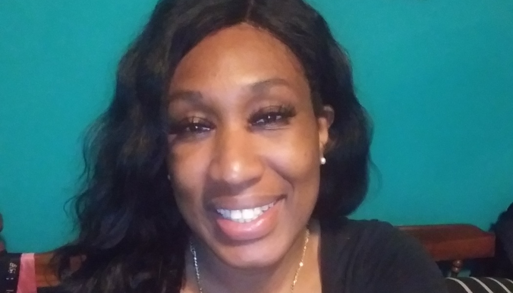 A smiling Black woman with long dark wavy hair against a bright teal wall.
