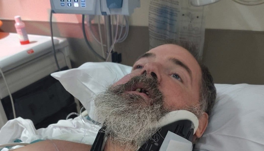 A bearded man wearing a neck brace lies in a hospital bed surrounded by medical equipment.