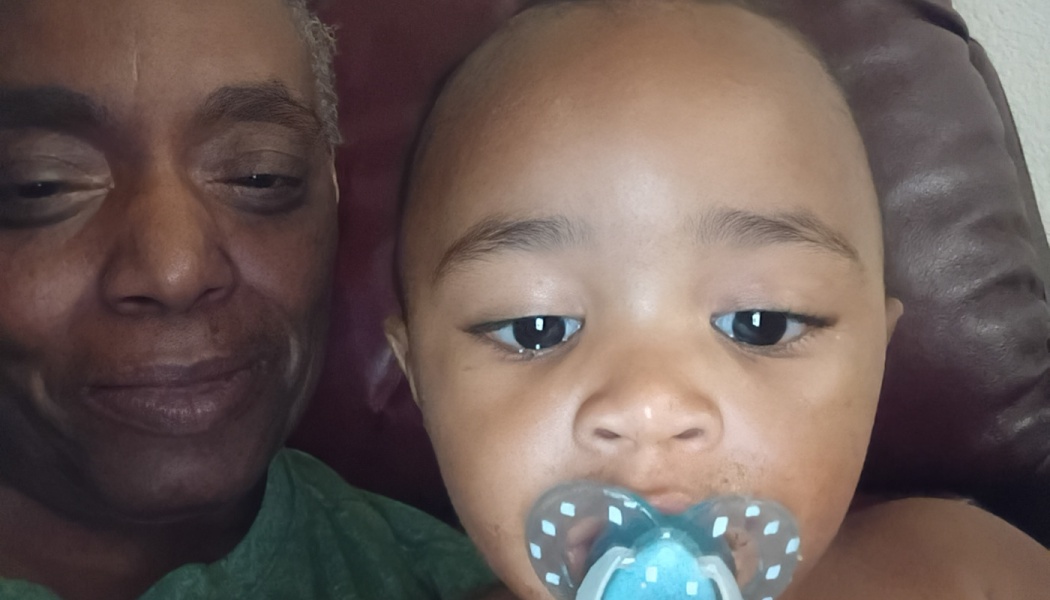 Close-up selfie of a smiling Black woman resting alongside a baby with a blue pacifier.