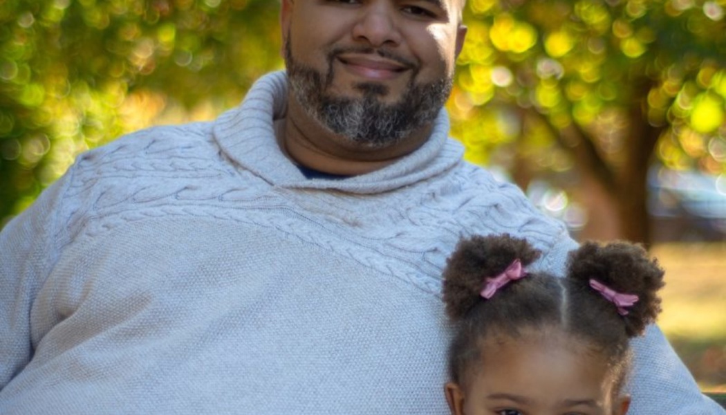 A smiling bearded man in a grey sweater sits outdoors with his young daughter wearing pink hair bows.