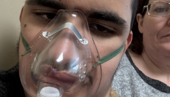 Close-up of a young man wearing a clear oxygen mask, with a woman in glasses sitting beside him.